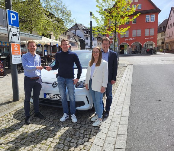 Vier Personen stehen auf einem Platz in einer Innenstadt vor einem weißen VW E-Auto. Zwei Männer links geben sich symbolisch den Autoschlüssel in die Hand. Rechts daneben stehen eine Frau im weißen Blazer und ein weiterer Mann im dunklen Sakko. Im Hintergrund sind ein Straßencafé, ein „E-Carsharing“-Schild mit Stadtwerk-Logo, Fachwerkhäuser und ein rotes Gebäude mit der Aufschrift „Konditorei“ zu sehen. Es ist sonnig, die Bäume haben frisches Frühlingsgrün.