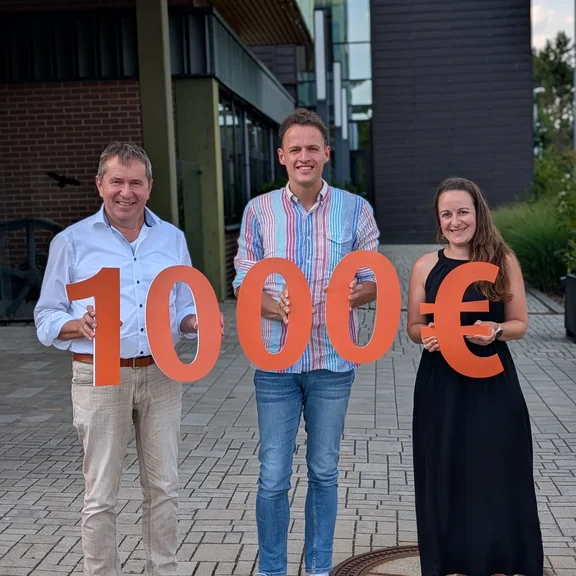 Spendenübergabe Lebenshilfe Drei Personen vor einem modernen Gebäude präsentieren einen symbolischen Spendenscheck in Höhe von 10.000 Euro, dargestellt durch große orangefarbene Zahlen und ein Eurozeichen.