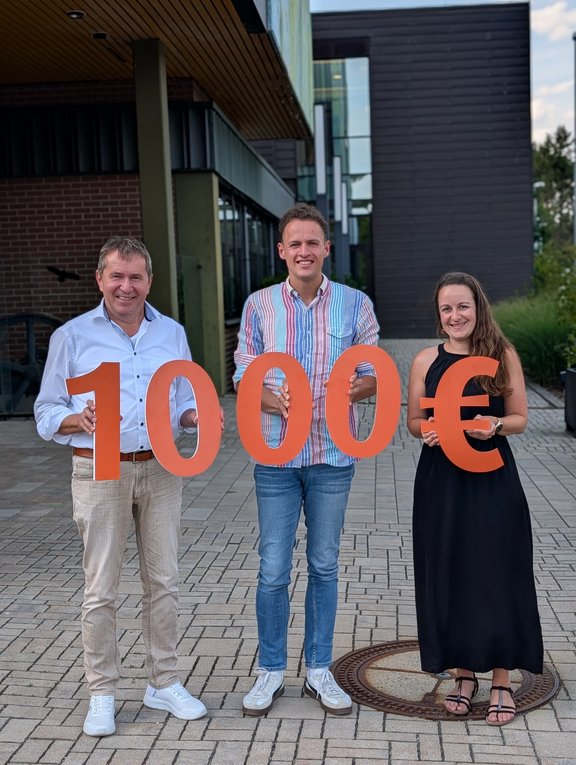 Drei Personen vor einem modernen Gebäude präsentieren einen symbolischen Spendenscheck in Höhe von 10.000 Euro, dargestellt durch große orangefarbene Zahlen und ein Eurozeichen.