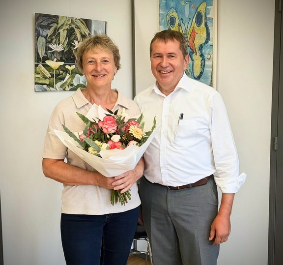 Mitarbeiterjubiläum Eine Person hält einen Blumenstrauß mit rosa und weißen Blumen in den Händen und steht gemeinsam mit einer weiteren Person in einem Büro. Beide blicken in die Kamera. Im Hintergrund sind zwei gerahmte Kunstwerke an einer hellen Wand zu sehen. Die Aufnahme entstand anlässlich eines Mitarbeiterjubiläums.