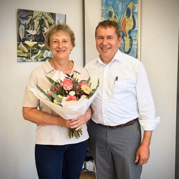Mitarbeiterjubiläum Eine Person hält einen Blumenstrauß mit rosa und weißen Blumen in den Händen und steht gemeinsam mit einer weiteren Person in einem Büro. Beide blicken in die Kamera. Im Hintergrund sind zwei gerahmte Kunstwerke an einer hellen Wand zu sehen. Die Aufnahme entstand anlässlich eines Mitarbeiterjubiläums.