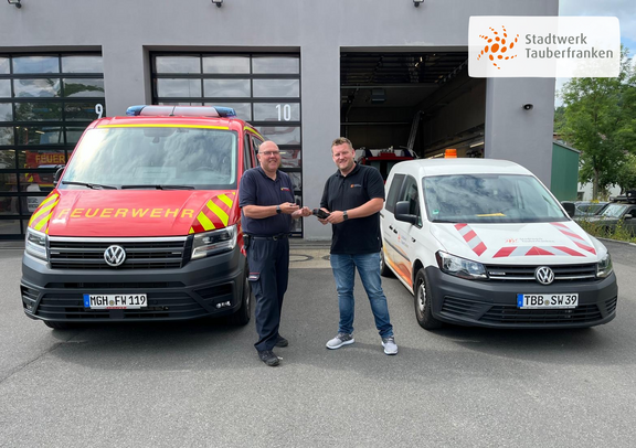 Zwei Männer stehen vor einem Feuerwehrgebäude und reichen sich lächelnd ein kleines schwarzes Gerät. Links steht ein Feuerwehrmann in Uniform vor einem roten Einsatzfahrzeug mit der Aufschrift "FEUERWEHR". Rechts steht ein Mitarbeiter des Stadtwerks Tauberfranken vor einem weißen Einsatzfahrzeug mit orangen Markierungen und dem Stadtwerk-Logo. Im Hintergrund sieht man offene Tore mit weiteren Fahrzeugen.