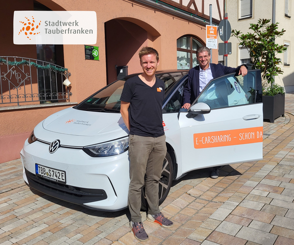 Zwei Männer stehen neben einem weißen VW ID.3 mit dem Logo des Stadtwerks Tauberfranken. Der Mann links trägt ein schwarzes T-Shirt mit Firmenlogo und lächelt in die Kamera. Der Mann rechts lehnt lächelnd an der geöffneten Fahrertür, trägt ein weißes Hemd mit dunklem Sakko. Im Hintergrund ist eine Hausfassade mit Fachwerkdetails und ein Carsharing-Hinweisschild zu sehen.