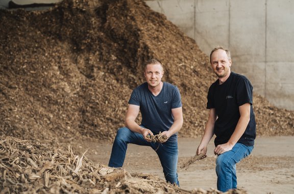 Zwei Männer stehen in Jeans und T-Shirt gekleidet in einer Halle in der Hackschnitzel gelagert werden. Sie lächeln in die Kamera und halten Holzhackschnitzel in der Hand.