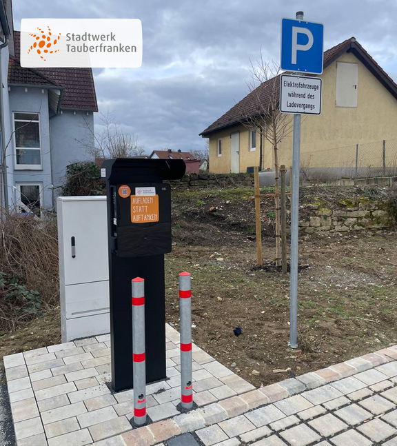 Ladestation der Stadtwerk Tauberfranken mit dem Slogan „Aufladen statt Auftanken“, aufgestellt auf gepflastertem Boden mit zwei rot-grauen Absperrpfosten. Daneben ein Schild mit Hinweis: „Elektrofahrzeuge während des Ladevorgangs“. Im Hintergrund Wohnhäuser und ein trüber Himmel.