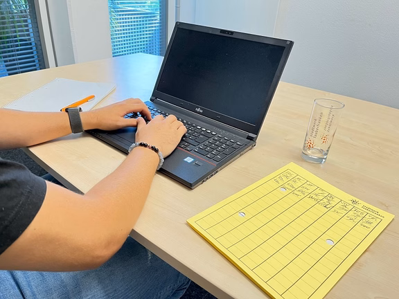 Energiesparen Person arbeitet an einem Laptop auf einem Holztisch, daneben liegt eine gelbe Mappe. Moderne Büroumgebung mit Fenster im Hintergrund.