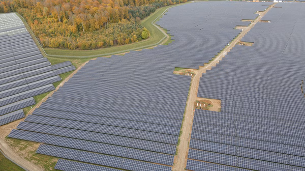 Luftaufnahme eines riesigen Solarparks mit unzähligen Solarpanelen, die sich über eine weite Fläche erstrecken. Ein sandiger Weg verläuft durch die Mitte der Anlage. Die Umgebung besteht aus grünen Wiesen und herbstlich gefärbten Wäldern mit gelben, orangen und roten Blättern.