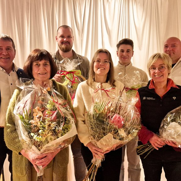 7 Personen stehen in schicker Kleidung vor einem weißen Vorhang. Vorne 3 Frauen mit einem Blumenstrauß in der Hand. Hinten 4 Männer. Die beiden in der Mitte halten einen großen Präsentekorb. Das Bild vermittelt die Situation einer Ehrung oder Beglückwünschung.