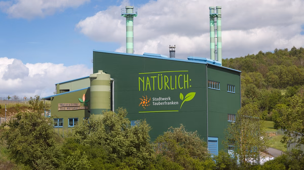 Grünes Industriegebäude mit der Aufschrift 'NATÜRLICH: Stadtwerk Tauberfranken' inmitten einer grünen Landschaft. Zwei grün-weiß gestreifte Schornsteine ragen in den Himmel. Auf der linken Seite ist ein weiteres Schild mit der Aufschrift 'Naturwärme Bad Mergentheim' zu sehen. Das Gebäude ist von Bäumen und Sträuchern umgeben, während der Himmel mit weißen Wolken bedeckt ist.