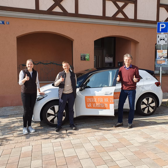Eine Frau und zwei Männer stehen vor uns neben einem Auto mit Stadtwerk Tauberfranken - Branding und lächeln in die Kamera.