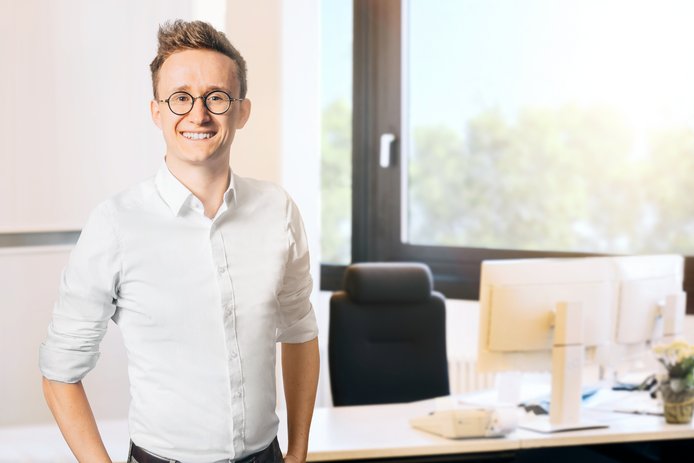 Eine Person mit Brille und weißem Hemd steht in einem hellen Büro. Im Hintergrund sind ein Schreibtisch mit mehreren Monitoren, ein Bürostuhl sowie ein großes Fenster mit Blick ins Grüne zu sehen.