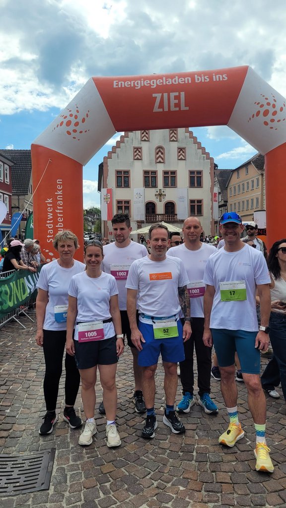 Mehrere Teilnehmende in weißen T‑Shirts und Laufschuhen stehen nach einem Lauf unter dem Zielbogen „Energieladen bis ins Ziel“ des Stadtwerks Tauberfranken, im Hintergrund ein historisches Rathaus auf einem Marktplatz.