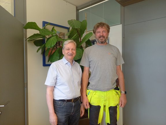 Zwei Männer stehen nebeneinander in einem Büro. Der Mann links trägt ein hellblaues Hemd und Jeans, der Mann rechts ein graues T-Shirt mit Logo des Stadtwerks Tauberfranken sowie gelb-schwarze Arbeitshose. Beide lächeln freundlich in die Kamera. Im Hintergrund befindet sich eine große grüne Zimmerpflanze sowie ein gerahmtes Bild an der Wand.