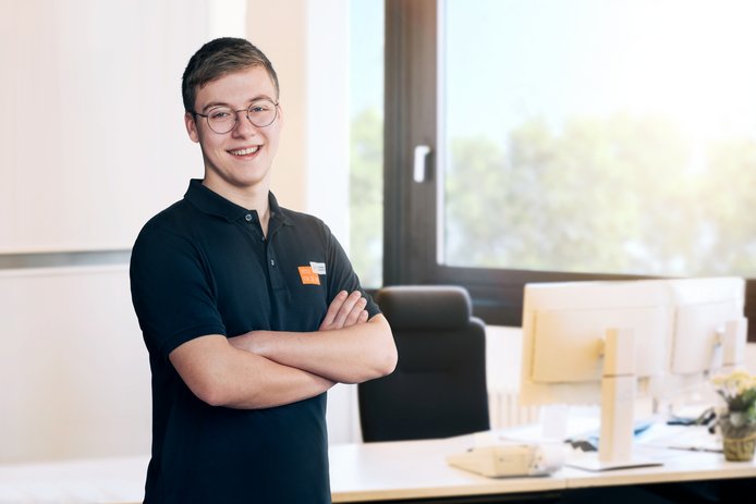 Mitarbeitender des Stadtwerks Tauberfranken steht in einem Büro mit verschränkten Armen vor einem Schreibtisch mit Monitoren. Im Hintergrund sind ein Fenster und unscharfe Arbeitsumgebung zu sehen.