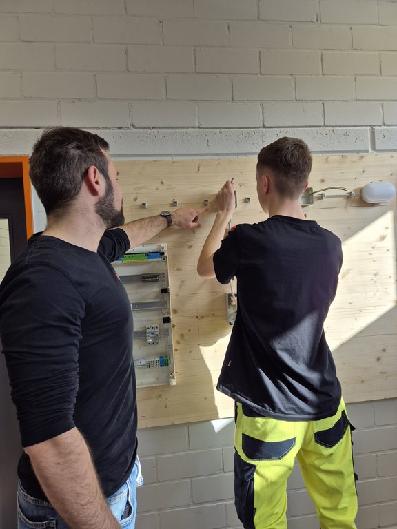 Ausbilder erklärt einem Auszubildenden den Aufbau einer elektrischen Schaltung an einer Übungswand in der Azubiwerkstatt.