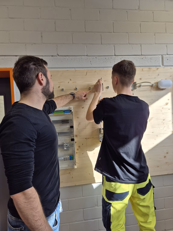 Ausbilder erklärt einem Auszubildenden den Aufbau einer elektrischen Schaltung an einer Übungswand in der Azubiwerkstatt.