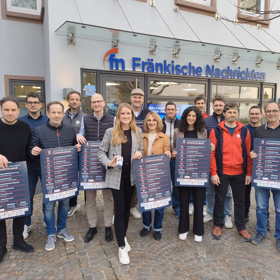 Gruppe von Menschen steht vor dem Gebäude der „Fränkische Nachrichten“ und hält Plakate zur Volkslaufserie der Laufgemeinschaft Tauberfranken in die Kamera; Außenaufnahme auf einem gepflasterten Platz.