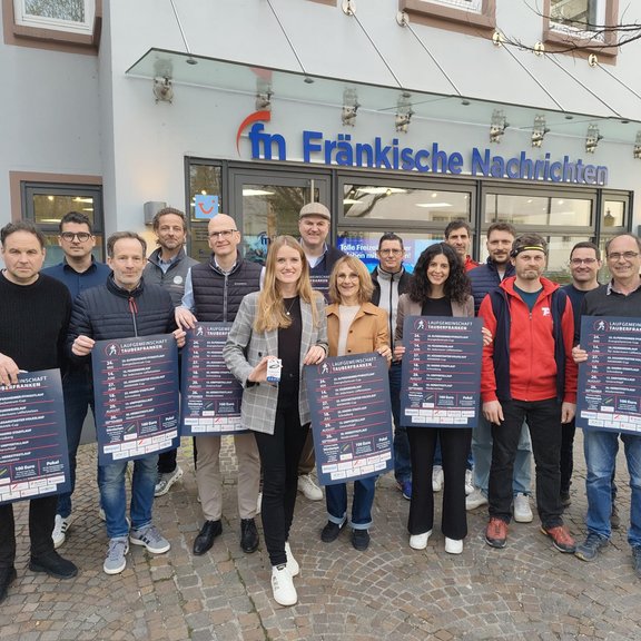 Gruppe von Menschen steht vor dem Gebäude der „Fränkische Nachrichten“ und hält Plakate zur Volkslaufserie der Laufgemeinschaft Tauberfranken in die Kamera; Außenaufnahme auf einem gepflasterten Platz.