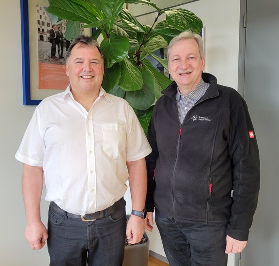 Betriebsjubiläum Zwei lächelnde Männer stehen nebeneinander in einem Büro. Einer trägt ein weißes Hemd, der andere eine schwarze Fleecejacke mit dem Logo des Stadtwerks Tauberfranken. Im Hintergrund befindet sich eine große grüne Pflanze und ein gerahmtes Poster an der Wand.