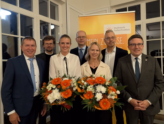 Infoveranstaltung Es zeigt eine Gruppe von sieben Personen bei einer Veranstaltung, zwei davon mit großen Blumensträußen. Im Hintergrund ist ein Banner mit dem gut erkennbaren Schriftzug „Stadtwerk Tauberfranken“ zu sehen.