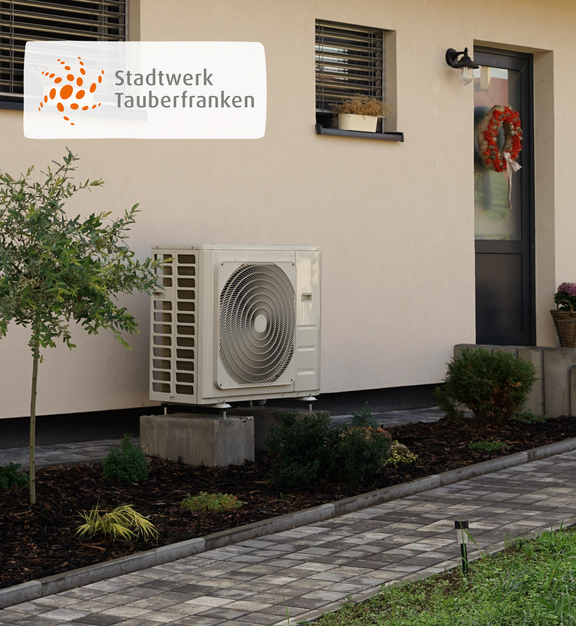 Steuerbare Verbrauchseinrichtung Außeneinheit einer Wärmepumpe vor einem modernen Wohnhaus mit Vorgarten und gepflastertem Weg. In der oberen linken Ecke befindet sich das Logo des Stadtwerks Tauberfranken.