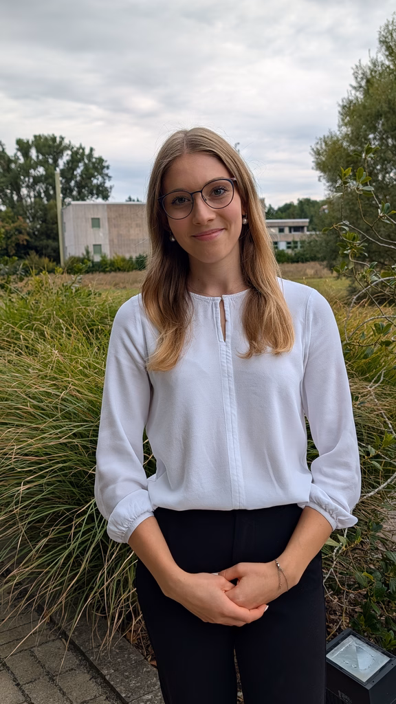 Mia Schiller orträtaufnahme einer jungen Mitarbeiterin mit Brille und heller Bluse. Die Person steht im Freien vor einer Grünanlage mit Bäumen und Gebäuden im Hintergrund.
