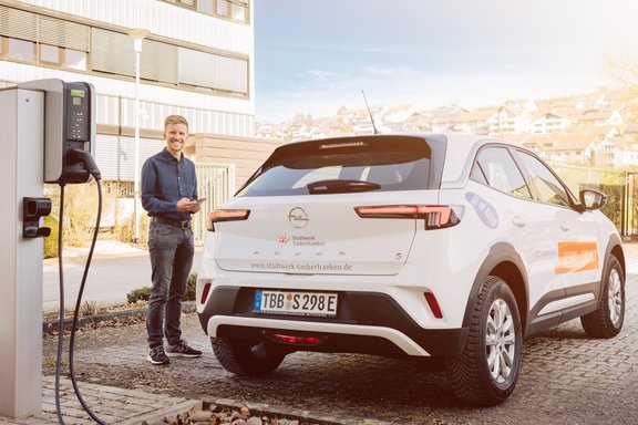 Mann mit kurzem blondem Haar und blauem Hemd steht lächelnd links neben einem weißen Opel SUV mit orangefarbenem Logo. Links im Bild ist eine Ladesäule sichtbar.