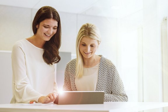2 Frauen stehen vor einem Laptop auf einem Stehtisch nebeneinander und unterhalten sich