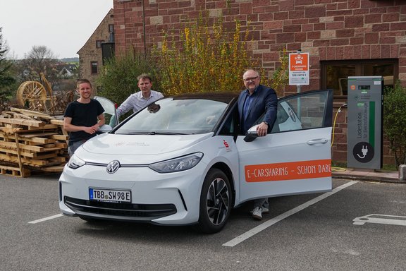 Drei Männer stehen neben einem weißen Elektroauto mit der Aufschrift „E-Carsharing – Schon dabei!“ vor einer Ladesäule der Stadtwerk Tauberfranken. Das Auto ist an die Ladesäule angeschlossen. Im Hintergrund sind eine rustikale Mauer, Holzstapel und ein Wasserrad zu sehen.