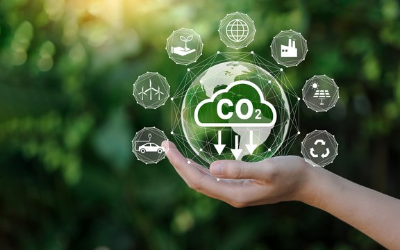 Eine Hand hält ein digitales Symbol mit einer CO₂-Wolke und Pfeilen, die auf Reduktion hinweisen. Rundherum sind Icons zu Nachhaltigkeit, wie E-Auto, Recycling, Solarenergie, Windkraft und Pflanzen. Der Hintergrund ist grün und unscharf – Symbolbild für Klimaschutz und CO₂-Reduktion.