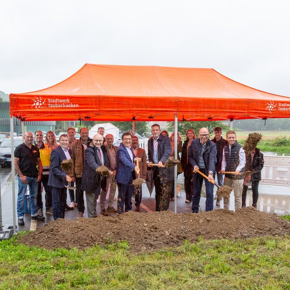 Das Bild vermittelt den Eindruck eines Spatenstichs für ein Projekt. Unter einem orangenen Pavillion mit dem Logo des Stadtwerks Tauberfranken stehen viele Menschen. Im Vordergrund stehen 6 Männer mit einem Spaten vor einem Erdhügel und werfen Erde in die Luft. Die Männer tragen Anzug und es regnet.