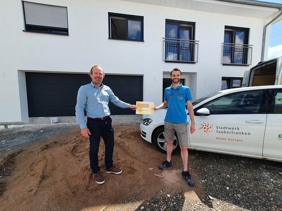 Mitarbeiter übergibt Gutschein an Kunde, im Hintergrund ist ein Stadtwer-Auto vor eine weißen Neubau-Einfamilienhaus