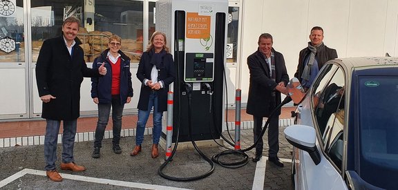Verantwrtlihe stehen vor einer Schnellladesäule und einer stöpselt das Auto davor gerade an