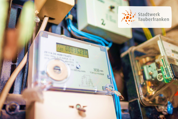 Smart Meter Nahaufnahme eines Smart Meters mit sichtbarem Zählerstand, umgeben von elektrischen Leitungen und weiteren Bauteilen. Rechts oben ist das Logo von Stadtwerk Tauberfranken eingeblendet.