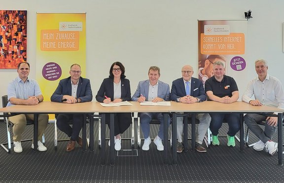 Sieben Personen sitzen an einem Tisch bei der Unterzeichnung einer Kooperationsvereinbarung. Im Hintergrund stehen zwei Roll-up-Banner des Stadtwerks Tauberfranken zu Energie- und Internetangeboten.