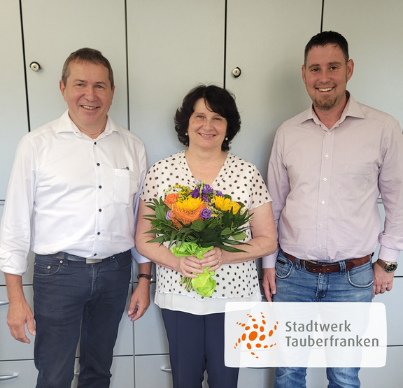 Betriebsjubiläum Drei Personen stehen vor einem grauen Aktenschrank und lächeln in die Kamera. In der Mitte hält eine Frau mit dunkelbraunen Haaren einen bunten Blumenstrauß mit Sonnenblumen, Rosen und anderen Blüten. Links und rechts von ihr stehen zwei Männer in Hemd und Jeans. Unten rechts im Bild ist das Logo von „Stadtwerk Tauberfranken“ eingeblendet.