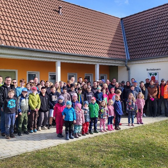 Viele Personen von jung bis alt stehen vor einem Gebäude und lächeln in die Kamera. Die Sonne scheint.