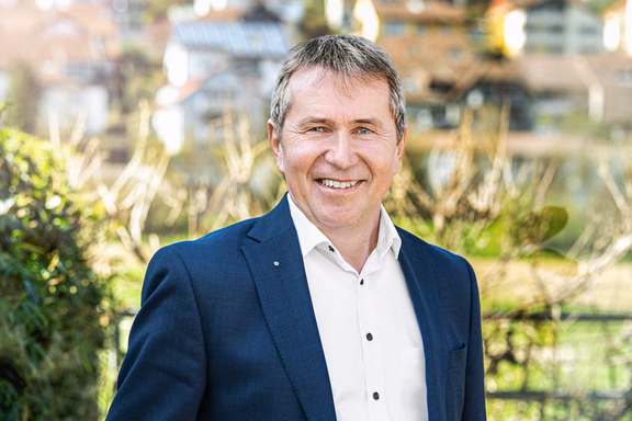 Ein Mann mittleren Alters steht lächelnd mit verschränkten Armen in einem modernen, hellen Bürogebäude. Er trägt einen dunklen Anzug mit weißem Hemd und eine elegante Armbanduhr. Die Sonne scheint durch große Fenster im Hintergrund und sorgt für eine warme Lichtstimmung.