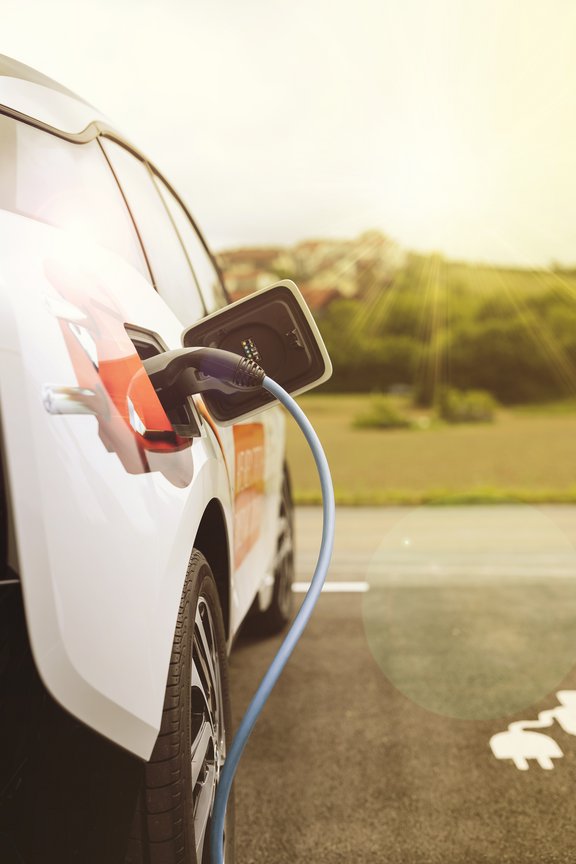 Ladevorgang Elektroauto mit Ladekabel, das an einer Ladestation angeschlossen ist, im Freien auf einem Parkplatz. Im Hintergrund ist ein Wohngebiet. Die Sonne scheint.