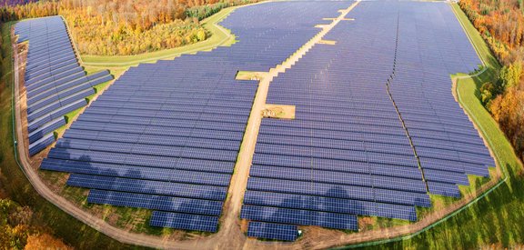 PV Gickelfeld Luftaufnahme eines riesigen Solarparks mit unzähligen Solarpanelen, die sich über eine weite Fläche erstrecken. Ein sandiger Weg verläuft durch die Mitte der Anlage. Die Umgebung besteht aus grünen Wiesen und herbstlich gefärbten Wäldern mit gelben, orangen und roten Blättern.