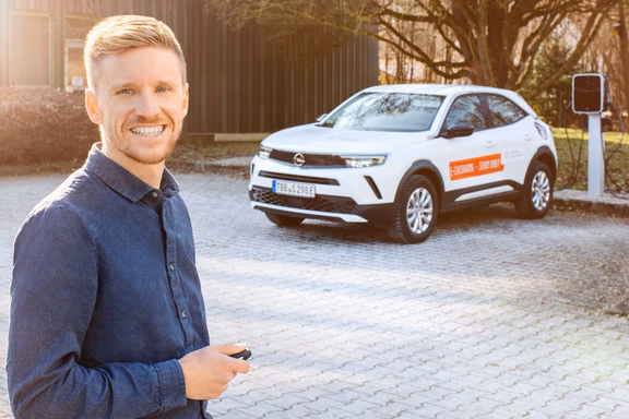 Elektromobilität Mann mit kurzem blondem Haar und blauem Hemd steht lächelnd vor einem weißen Opel SUV mit orangefarbenem Logo. Im Hintergrund ist eine Ladesäule sichtbar.