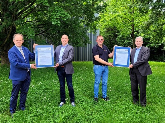 4 Verantwortliche stehen auf einer Wiese und je 2 halten einen großen Bilderrahmen hoch mit einer Urkunde