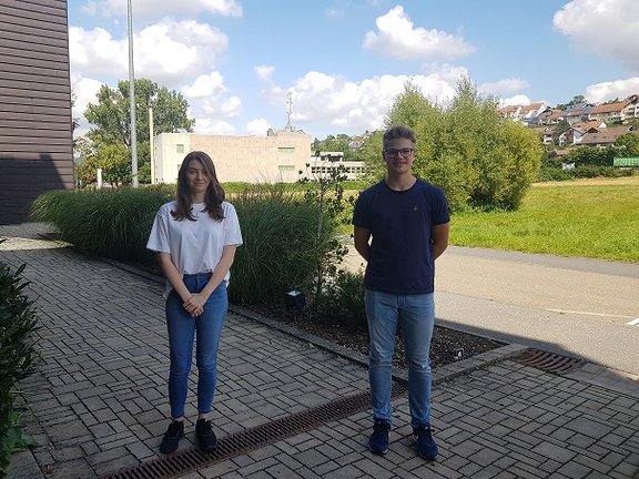 Eine Auszubildende und ein Auszubildender stehen nebeneinander vor einer kleinen Hecke