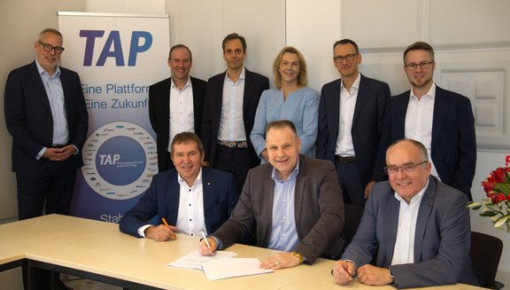 Männer und Frauen in Business-Kleidung sitzen und stehen an einem Tisch, halte teilweise einen Stift in der Hand. Hinten ist ein Roll-Up mit der Aufschrift "TAP" zu sehen. Die Szene vermittelt eine Vertragsunterzeichnung oder einen Abschluss.