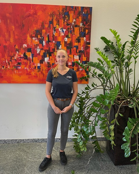 Mitarbeiterin steht vor organenem Bild und neben grüner Pflanze